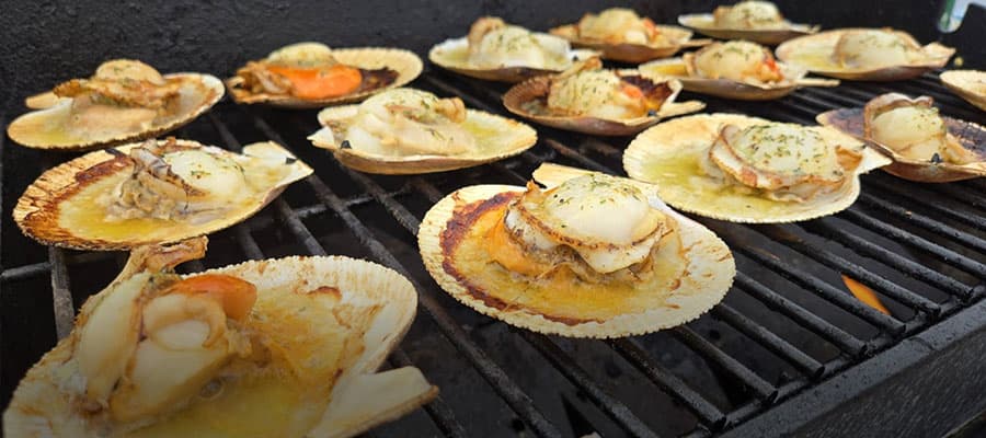 A pile of fresh cloudberries sits on a wooden cutting board. In the background, there's blurred imagery of green foliage and a wooden table. The cloudberries are vibrant orange and red, indicating ripeness.
