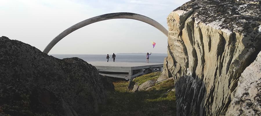 Three people stand near a large, curved stone sculpture overlooking the ocean. One person is flying a colorful kite. The scene is framed by rocky cliffs and grassy terrain.