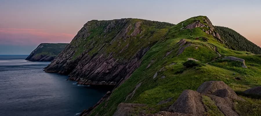 A scenic view of a rocky, green-clad cliff extending into the ocean at sunset. The sky is a gradient of pink and blue, with the sun casting a warm glow over the landscape. The rugged terrain is covered with grass and scattered rocks.