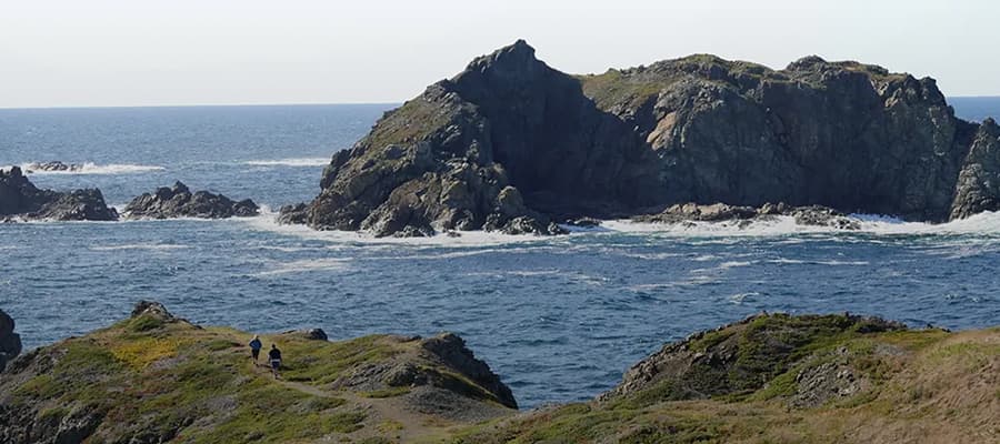 A rocky coastal scene with large formations jutting into the ocean under a clear sky. The sea is calm with gentle waves. In the foreground, two people walk along a narrow path bordered by grassy terrain.