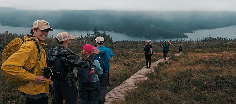 A group of hikers, wearing backpacks and caps, walk along a wooden path through grass and shrubs. A misty landscape with mountains and a body of water stretches out in the background under a cloudy sky.