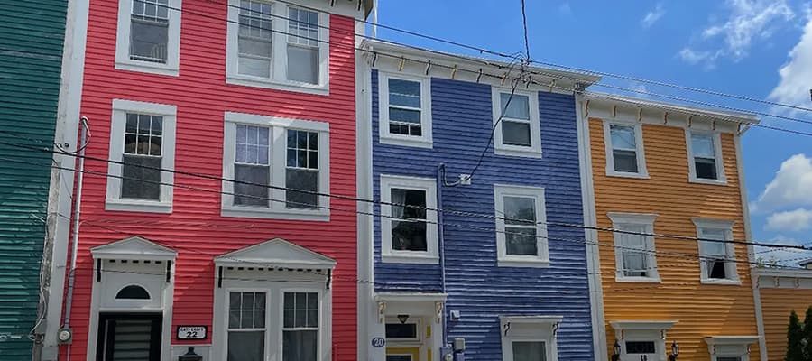 A row of colorful three-story houses with white trim. From left to right, the houses are green, red, blue, and yellow. The sky is partly cloudy, and power lines run across the scene.