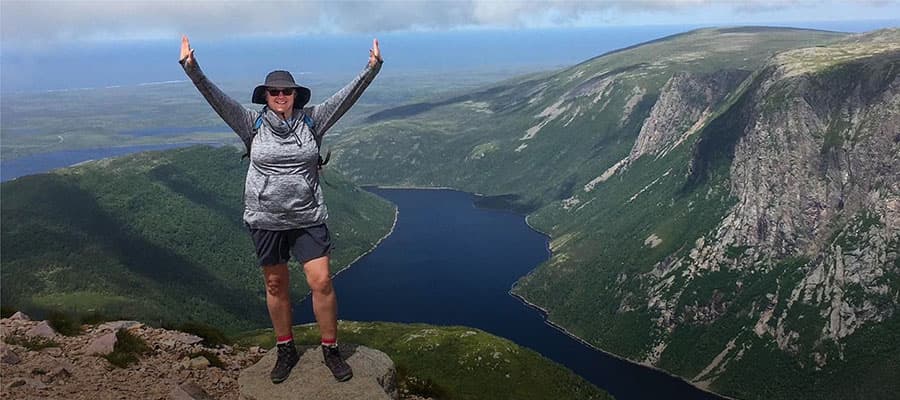 Three people stand near a large, curved stone sculpture overlooking the ocean. One person is flying a colorful kite. The scene is framed by rocky cliffs and grassy terrain.