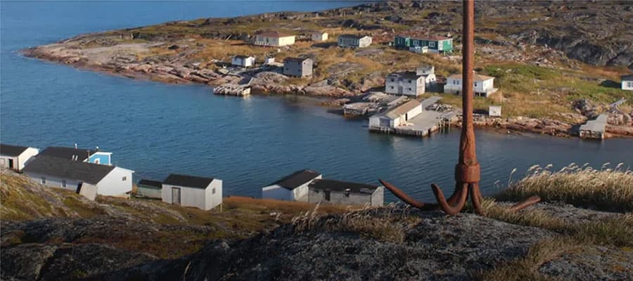 Two people walking on a grassy, rocky coastal landscape towards rocky cliffs. The ocean is visible on the right, with a clear blue sky and fluffy clouds above.