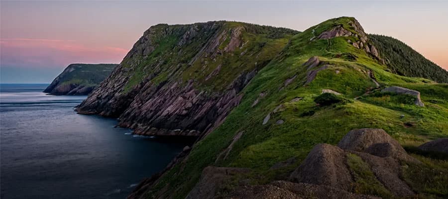 A scenic view of a rocky, green-clad cliff extending into the ocean at sunset. The sky is a gradient of pink and blue, with the sun casting a warm glow over the landscape. The rugged terrain is covered with grass and scattered rocks.