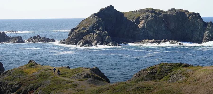 A rocky coastal scene with large formations jutting into the ocean under a clear sky. The sea is calm with gentle waves. In the foreground, two people walk along a narrow path bordered by grassy terrain.