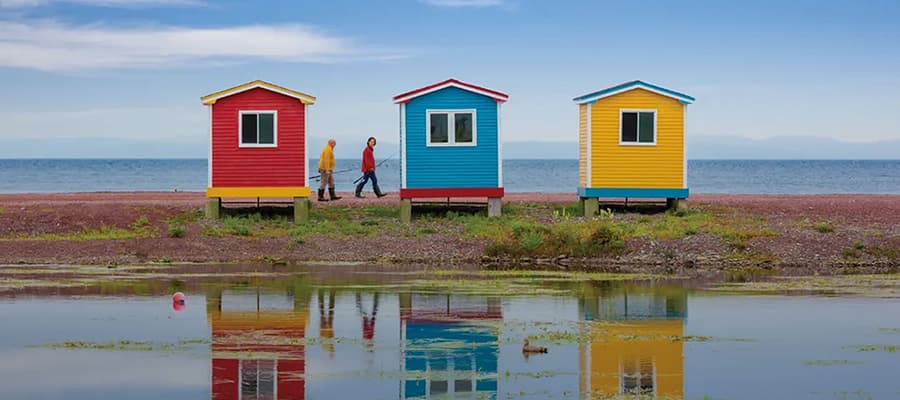 Three colorful beach huts—red, blue, and yellow—stand on a pebbled shore by the sea. Two people walk side by side, one in a yellow coat, the other in red. The huts and people are reflected in a still water surface.