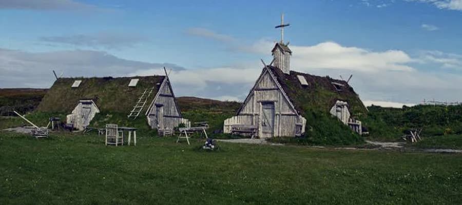 Viking-style sod-roofed buildings with wooden walls are set on a grassy landscape under a cloudy sky. A ladder leans against one building, and wooden furniture is scattered in front. The scene evokes a rustic, historical atmosphere.