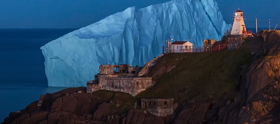 A large iceberg floats near the coastline, illuminated in blue hues. In the foreground, on a rocky hill, stands a white lighthouse beside several small buildings, under a dark blue sky.