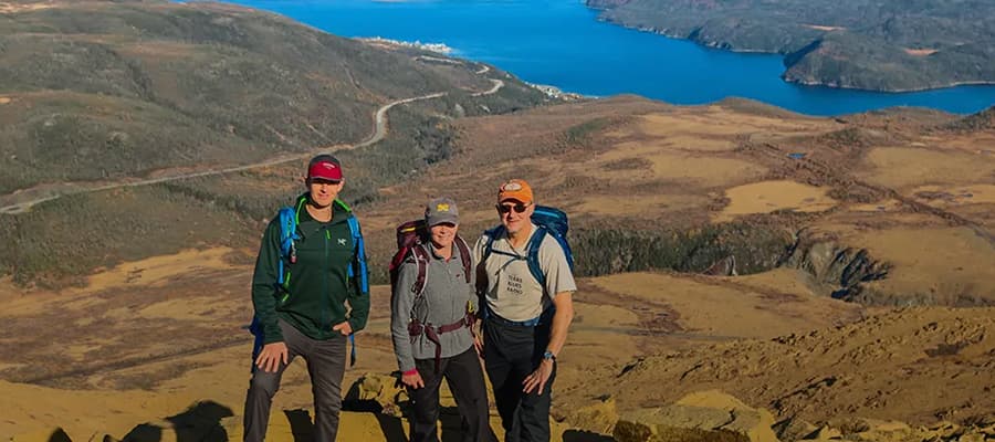 Three people stand on a rocky mountain top, wearing hiking gear and backpacks. A vast landscape with a winding road, a body of water, and rolling hills extends behind them under a clear blue sky.