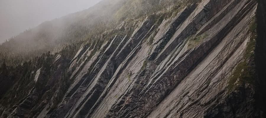 Steep mountain slope with jagged, layered rock formations and patches of greenery. A misty atmosphere covers the peak, creating a dramatic and rugged landscape.