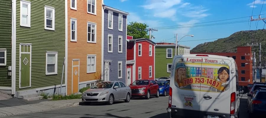A colorful row of houses in green, orange, red, and maroon along a hilly street with a Legend Tours van parked in front. The scene is under a clear blue sky.