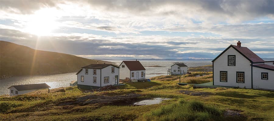 Three colorful beach huts—red, blue, and yellow—stand on a pebbled shore by the sea. Two people walk side by side, one in a yellow coat, the other in red. The huts and people are reflected in a still water surface.