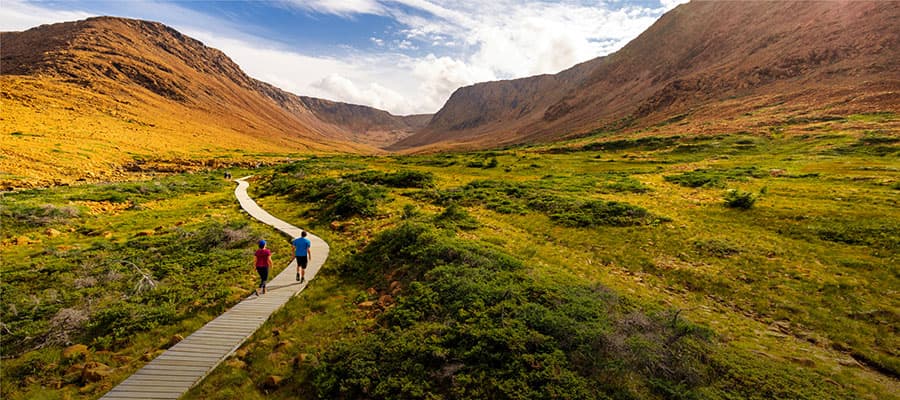 Three people stand on a rocky mountain top, wearing hiking gear and backpacks. A vast landscape with a winding road, a body of water, and rolling hills extends behind them under a clear blue sky.