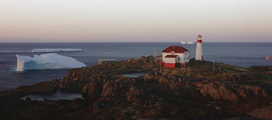 A solitary lighthouse and building sit on a rocky coastal landscape. An iceberg floats in the ocean nearby. The scene is under a pastel sky during sunset or sunrise, with calm waters reflecting soft colors. A small Canadian flag waves in the foreground.
