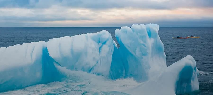 A large iceberg with jagged peaks floats in the ocean under a cloudy sky. A small kayak with two people is visible in the background, emphasizing the scale of the iceberg.
