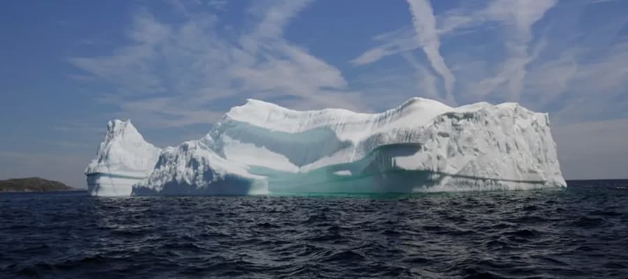 A large iceberg with a jagged, snowy surface floats in a dark blue sea under a sky with scattered clouds. The horizon features a small stretch of land. The iceberg has shades of white and light blue with some visible texture.