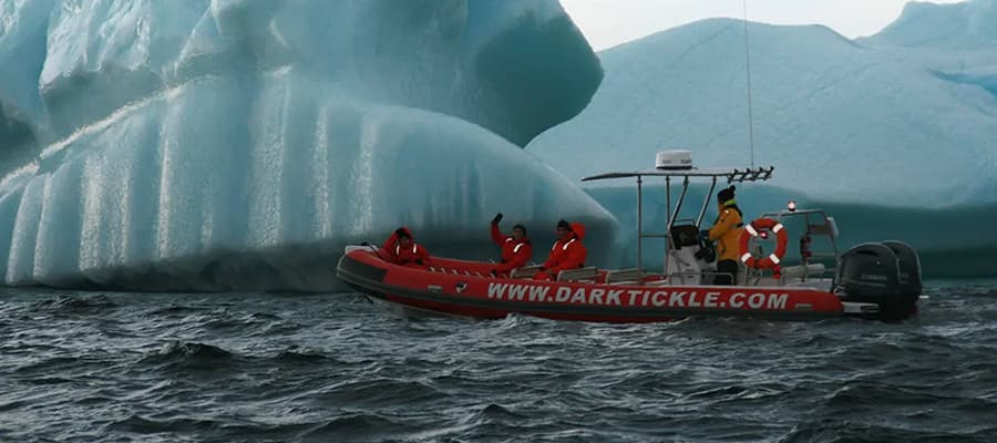 A group of people in red suits are on a red inflatable boat labeled "www.darktickle.com," navigating close to a large iceberg amidst dark ocean waters. One person is waving, and a lifebuoy is visible on the boat.