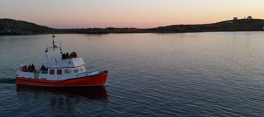 A red and white boat with people on board sails across calm waters at sunset. The sky is a gradient of orange and purple, and the shoreline with rocky terrain is visible in the background.