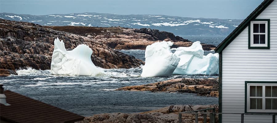 A solitary lighthouse and building sit on a rocky coastal landscape. An iceberg floats in the ocean nearby. The scene is under a pastel sky during sunset or sunrise, with calm waters reflecting soft colors. A small Canadian flag waves in the foreground.