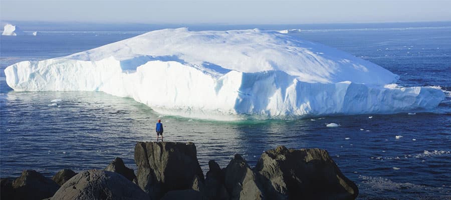 A large iceberg with jagged peaks floats in the ocean under a cloudy sky. A small kayak with two people is visible in the background, emphasizing the scale of the iceberg.