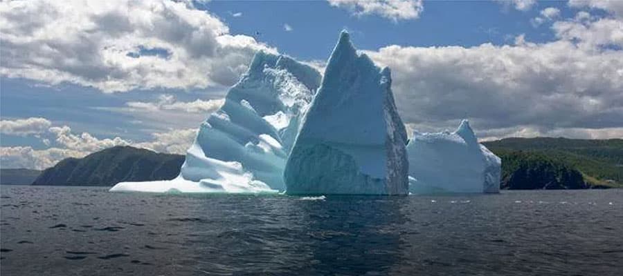 A large iceberg with a jagged, snowy surface floats in a dark blue sea under a sky with scattered clouds. The horizon features a small stretch of land. The iceberg has shades of white and light blue with some visible texture.
