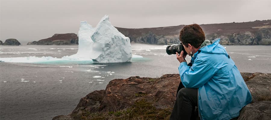 A group of people in red suits are on a red inflatable boat labeled "www.darktickle.com," navigating close to a large iceberg amidst dark ocean waters. One person is waving, and a lifebuoy is visible on the boat.