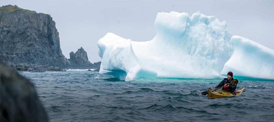 A red and white boat with people on board sails across calm waters at sunset. The sky is a gradient of orange and purple, and the shoreline with rocky terrain is visible in the background.