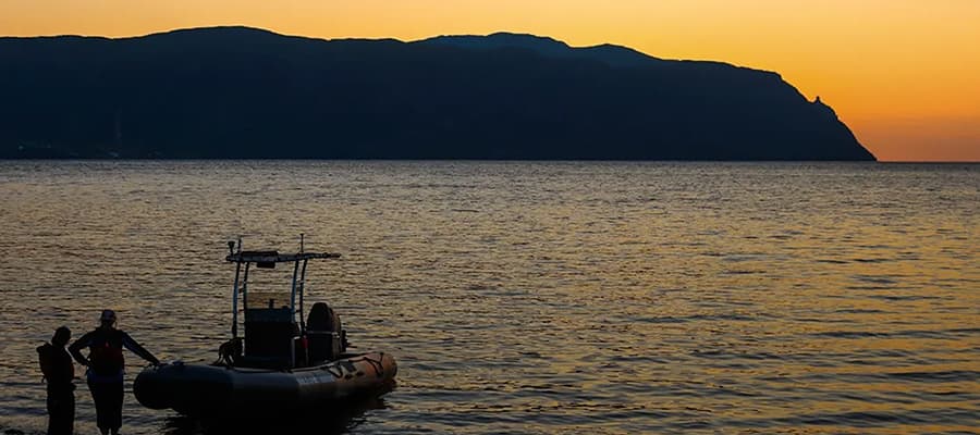 A small boat with two people stands near the shore at dusk, silhouetted against a calm sea. A mountainous coastline is visible in the background under the orange glow of the sunset sky.