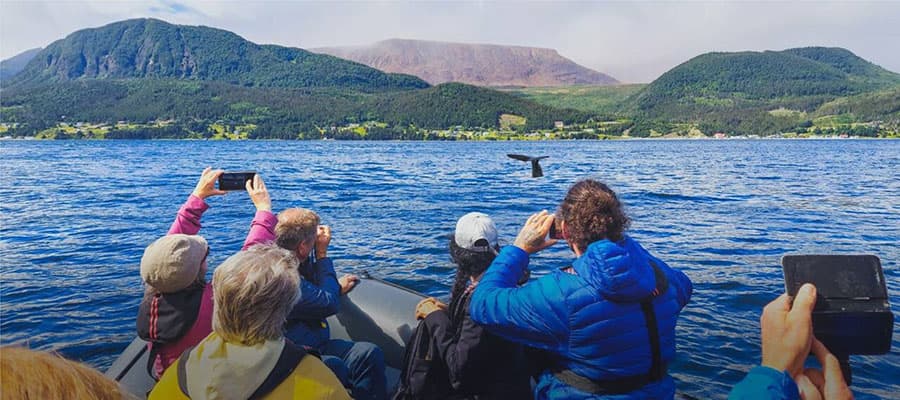 A small boat with two people stands near the shore at dusk, silhouetted against a calm sea. A mountainous coastline is visible in the background under the orange glow of the sunset sky.