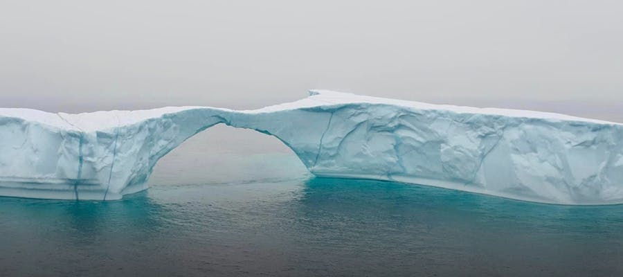 A long, skinny iceberg with an arch bridge sits in the Atlantic ocean.