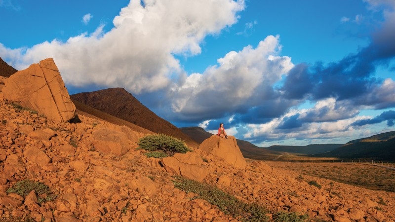 A traveller is resting on a boulder, surrounded by the red landscape of the Tablelands in Gros Morne National Park.