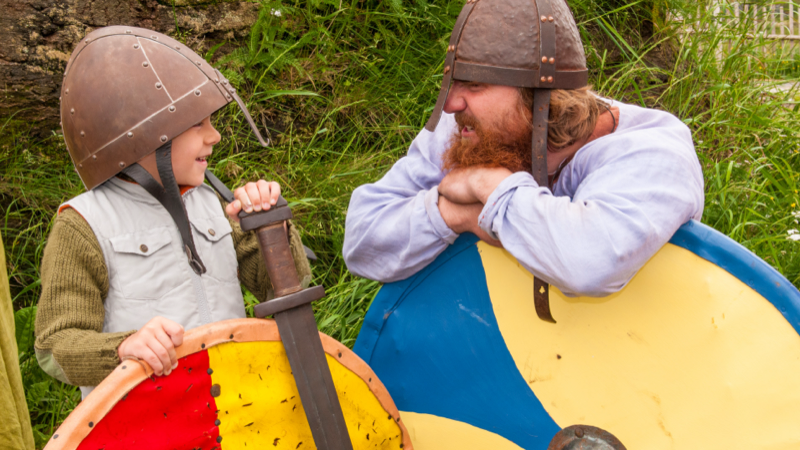 A reenactment actor is telling a fun story to a child. Both are wearing Viking outfits.