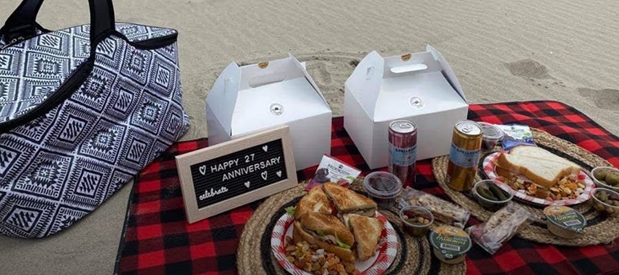 An assortment of food and drinks on a picnic blanket on the sand, with a sign that reads, “Happy 27 Anniversary.”
