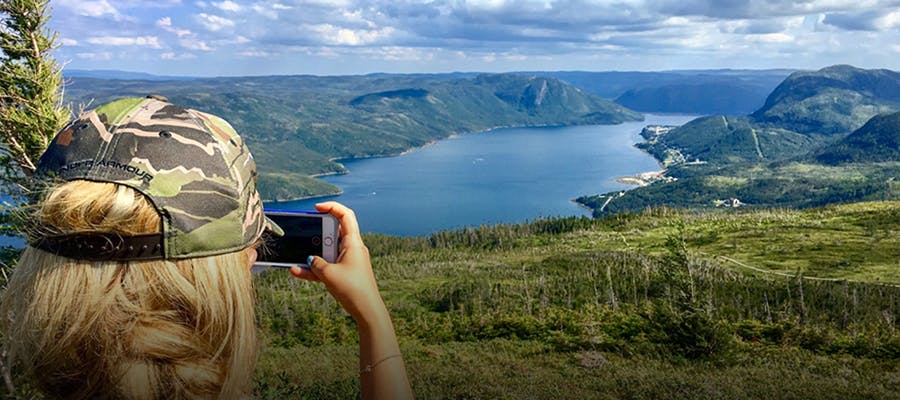 A traveller taking a photo with their phone of the view of the water and hills atop a Gros Morne National Park trail.