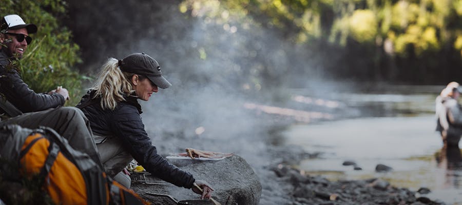 A woman cooking on a rocky beach by a river.