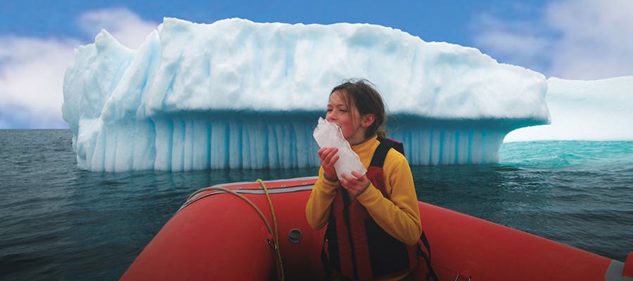A child sitting on a tour boat, licking a chunk of ice with an iceberg towering in the background.